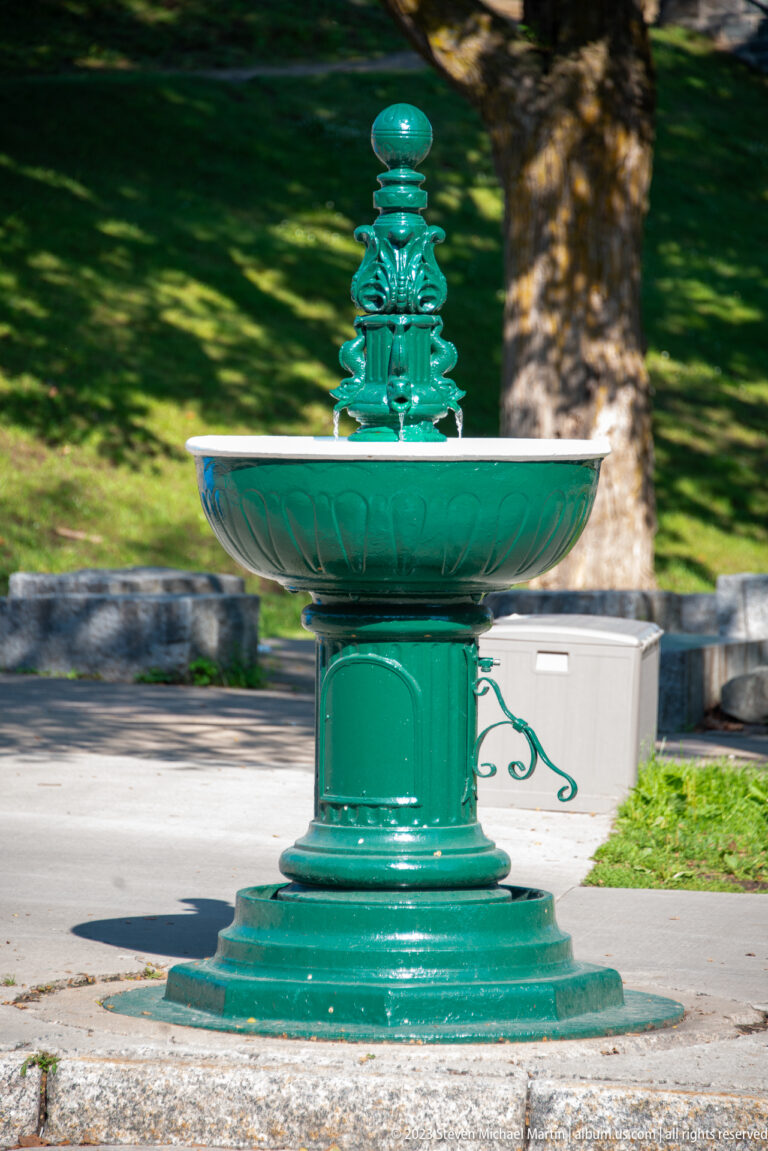 Green cast-metal public fountain with broad circular basin, decorative pedestal, embossed ornament, and finial beside a roadway.
