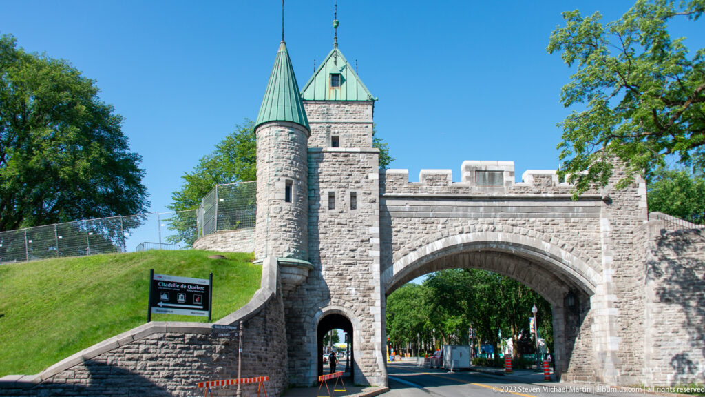 Front view of stone gateway with roadway arch, pedestrian opening, round turret, square tower, crenellations, and grassy rampart.