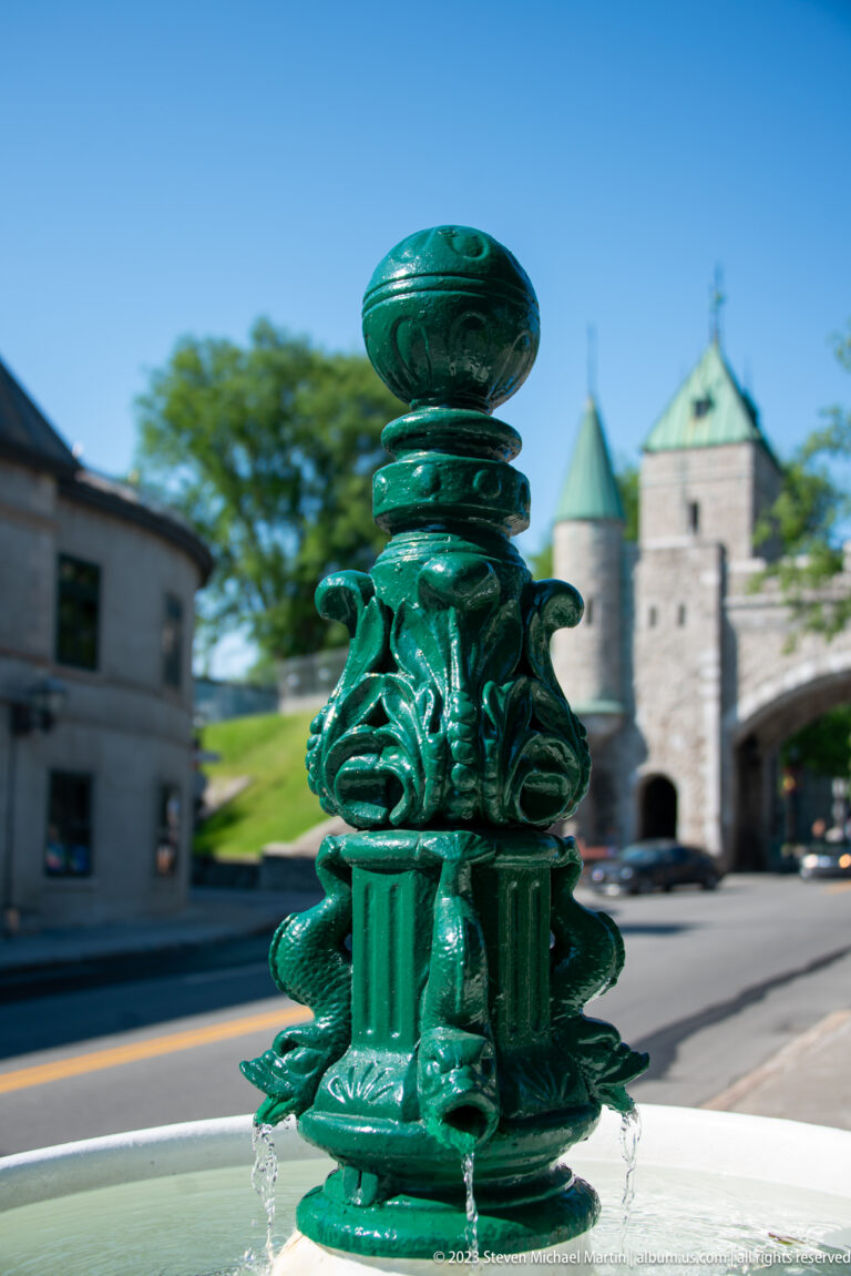Close detail of ornate green cast-metal fountain fittings and rounded finial, with a stone fortified gateway blurred behind.