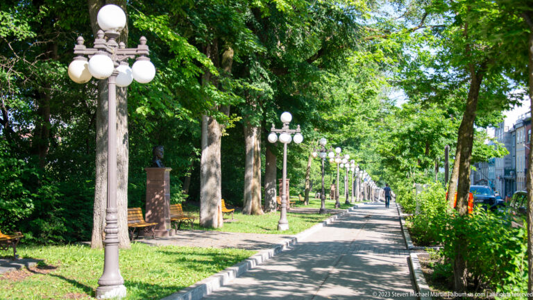 Long shaded pedestrian promenade lined with mature trees, benches, lawn, and repeating globe-topped streetlamps.