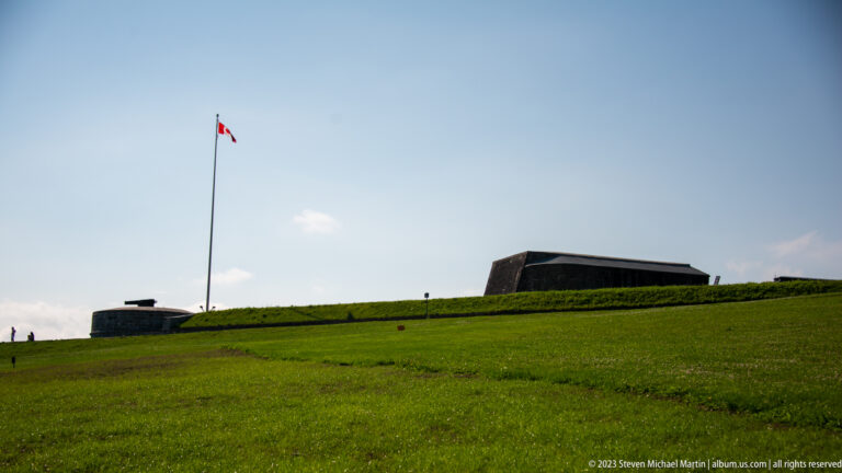 Grassy Citadelle earthworks with low angular fortification structures and a tall Canadian flagpole against blue sky.