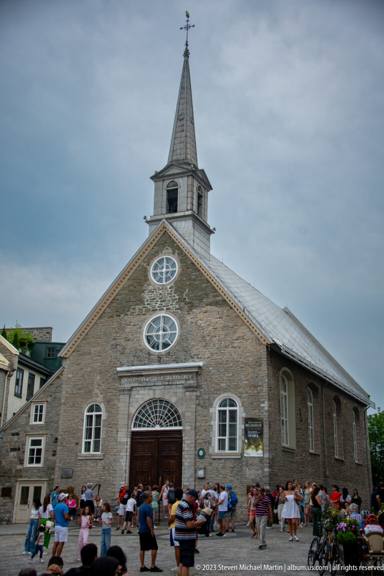 Front view of Notre-Dame-des-Victoires with stone façade, arched doorway, two circular windows, steep roof, spire, and crowd.