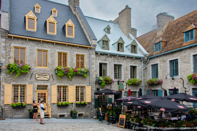 Stone houses with yellow and blue window trim and flower boxes above a busy restaurant terrace at Place Royale.