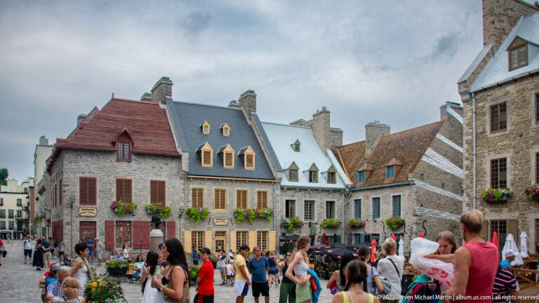 Crowds cross Place Royale before rows of stone houses with steep roofs, dormers, colorful trim, and flower boxes.