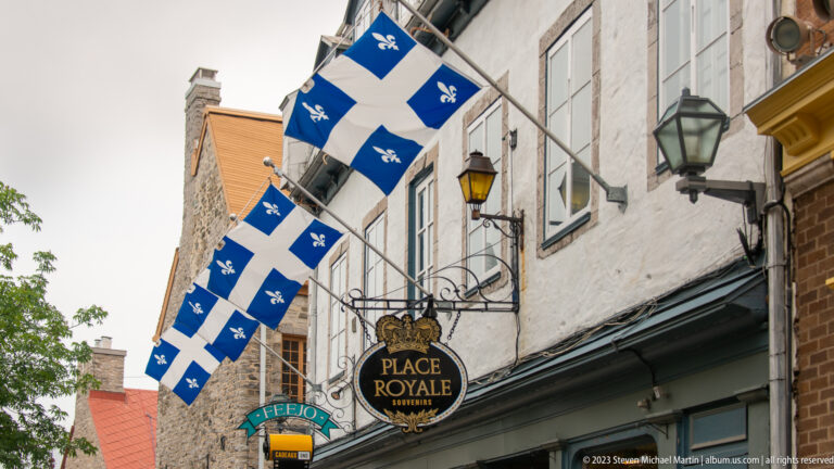 Several Quebec flags project over a narrow street above a hanging “Place Royale Souvenirs” sign and traditional lanterns.