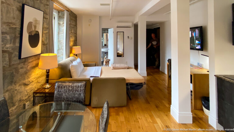 Hotel room with exposed stone walls, wood floor, seating area, bed, lamps, and a wall-mounted television.