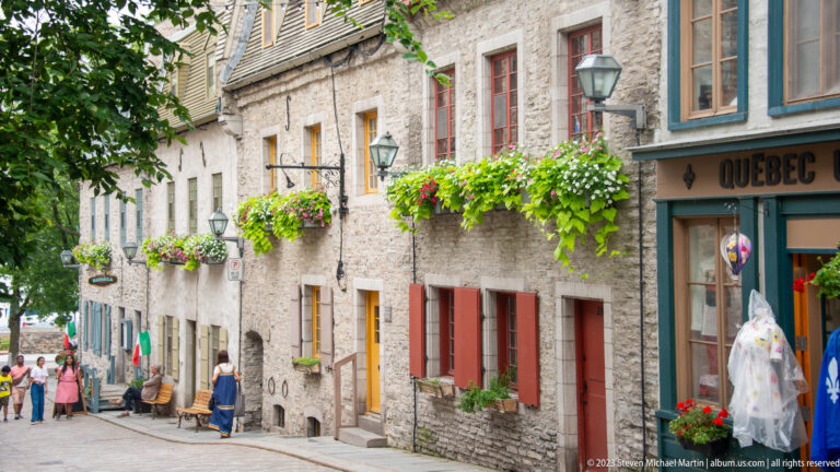 Narrow descending street with rough stone buildings, colorful window trim, hanging flower boxes, lanterns, and pedestrians.