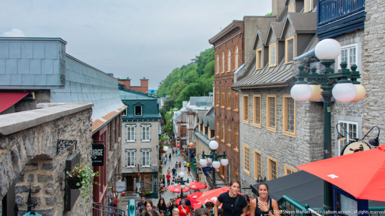 View down a steep Quebec City stairway lined with historic buildings, globe streetlamps, red umbrellas, and pedestrians.