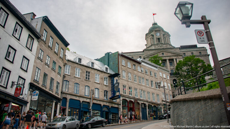 Quebec City Hall tower rises behind historic shops and parked cars along an Old Quebec street.