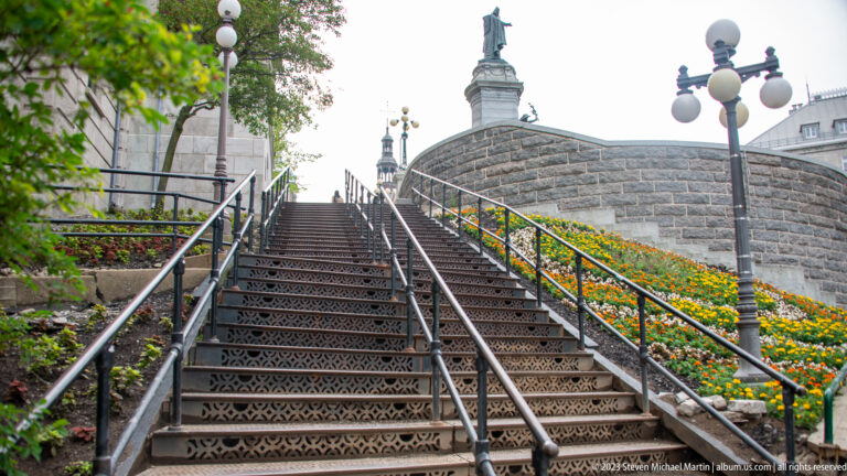 Decorative metal stairs rise beside a stone retaining wall planted with orange and yellow flowers, with globe lamps and a monument above.
