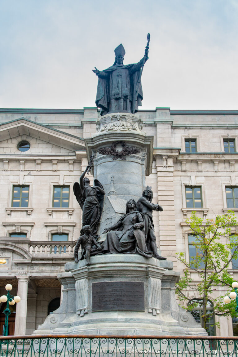 Bronze monument with a standing figure at the top and multiple sculpted figures around the stone base before a large building.