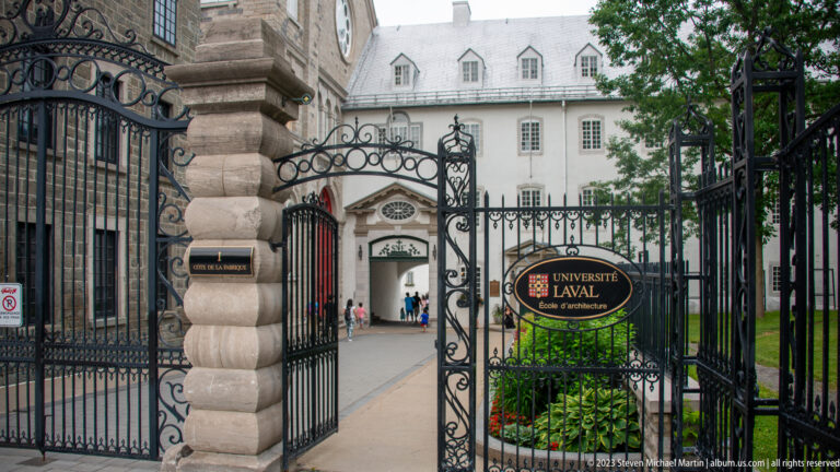 Wrought-iron gates and stone posts at the Université Laval École d’architecture entrance, with a pale historic building beyond.