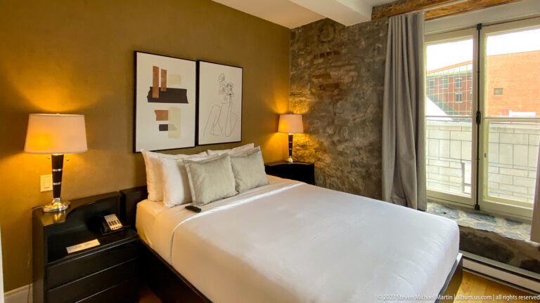 Made hotel bed beside an exposed stone wall, tall window with curtains, framed art, and two illuminated bedside lamps.