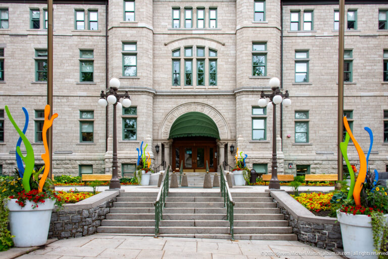 Symmetrical stone building with central arched entrance, broad stairs, globe lamps, flower beds, and colorful abstract sculptures.