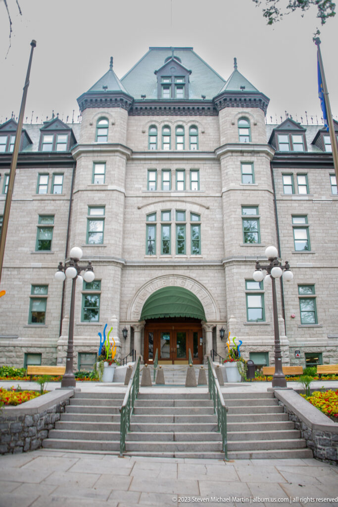 Large stone Quebec City Hall façade with steep roof, dormers, central arched entrance, broad steps, flags, and flower beds.