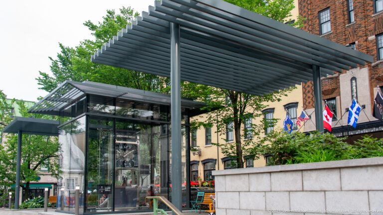 Glass pavilion beneath a dark metal slatted canopy, bordered by trees, stone walls, flags, and older masonry buildings.
