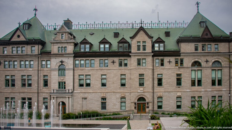 Long stone institutional building with gabled bays, green metal roof, many windows, and fountain jets across the foreground.