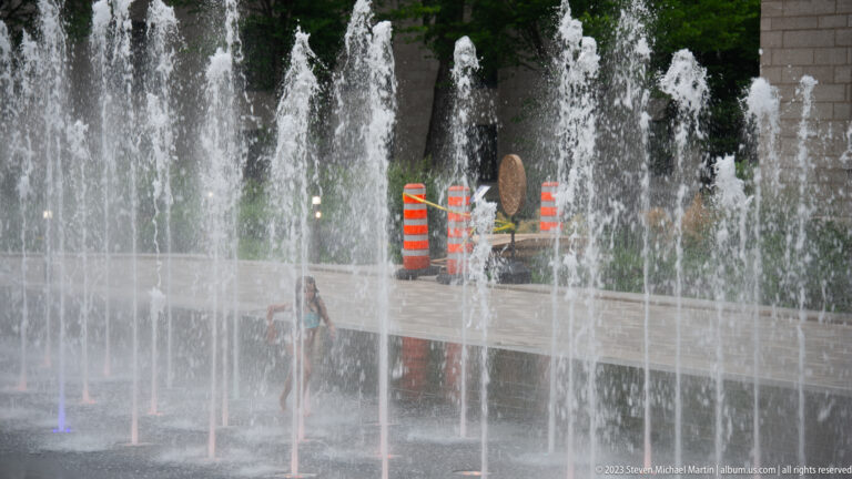 Rows of vertical fountain jets rise from pavement, partially obscuring a pedestrian and orange construction barrels behind them.