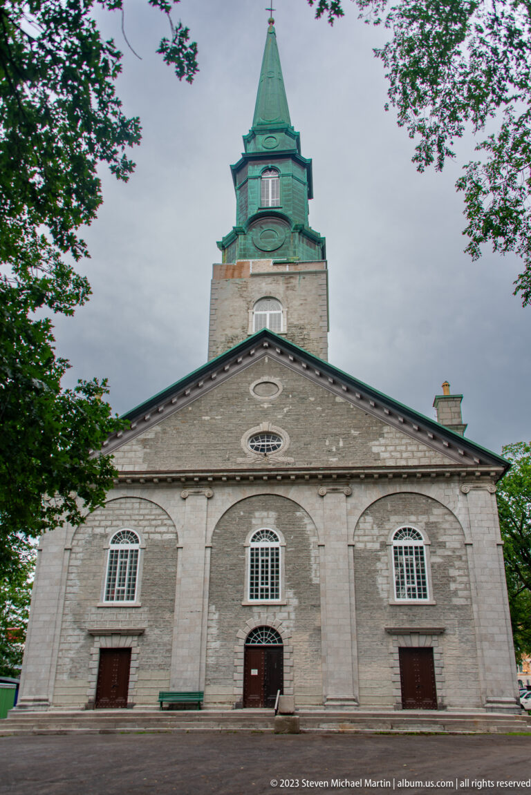 Symmetrical gray stone church with three doors, arched windows, triangular pediment, and tall central tower capped by a green spire.