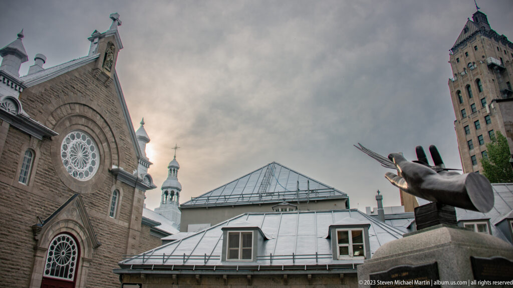 Stone church with rose window beside a dark sculpture and rooftops, with the tall Price Building rising at right under cloudy sky.