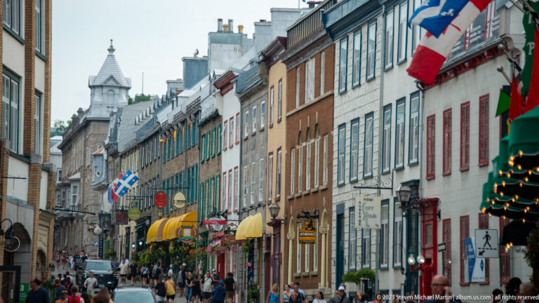 Crowded narrow street lined with colorful historic buildings, shop awnings, signs, Quebec flags, pedestrians, and vehicles.