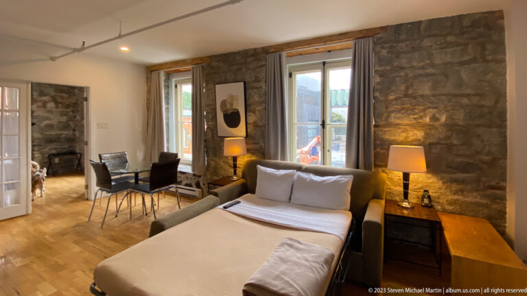 Bed in a stone-walled hotel room facing two chairs and tall windows, with lamps, artwork, and wood flooring.