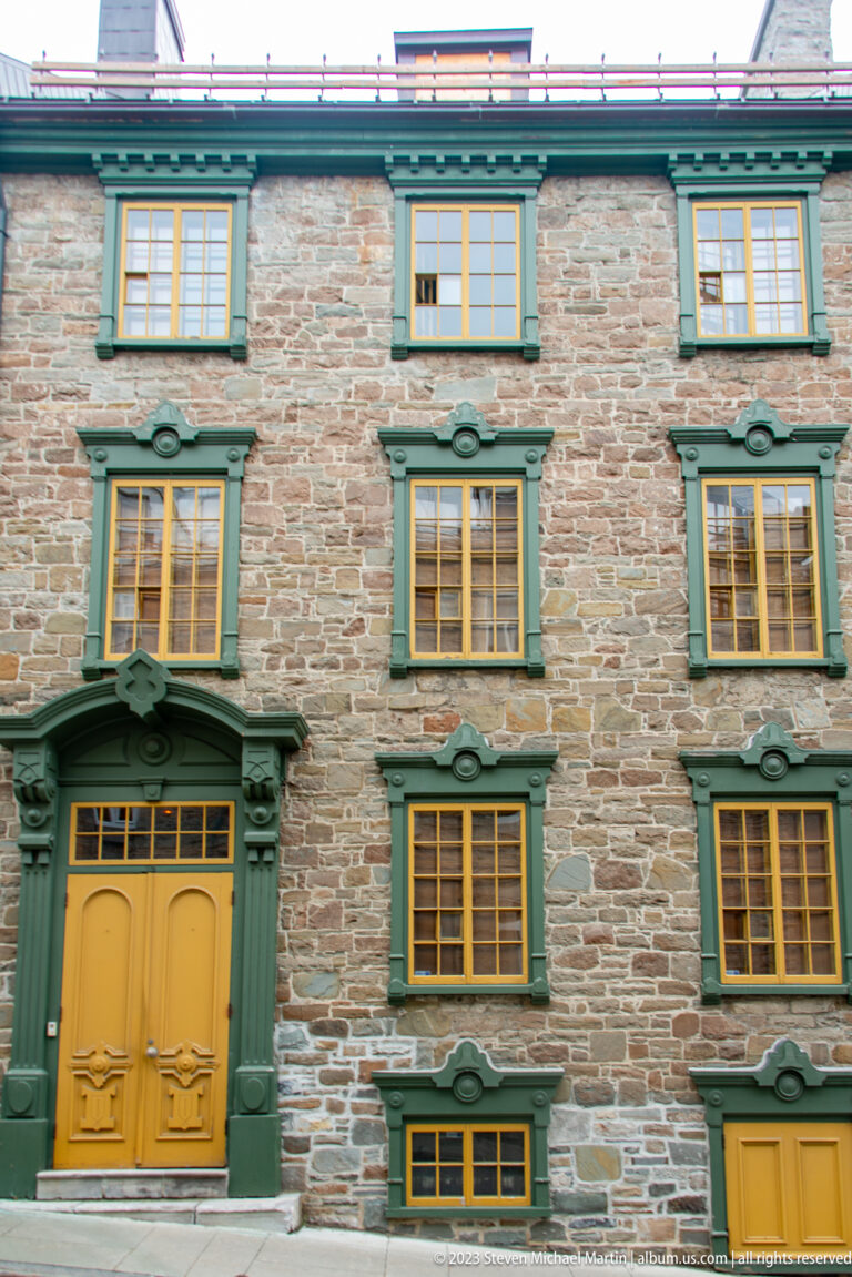 Stone townhouse façade with green window trim, small-paned windows, and ochre-yellow doors arranged in regular bays.