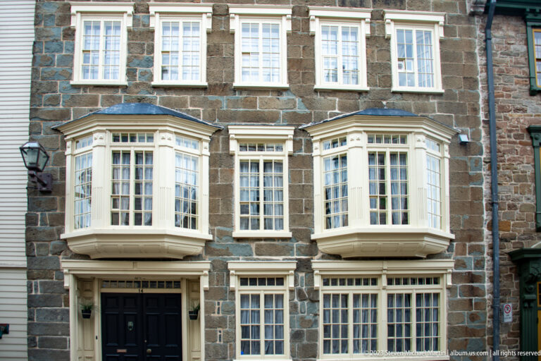 Dark stone townhouse with two white projecting bay windows, rows of multi-pane windows, and a black doorway below.