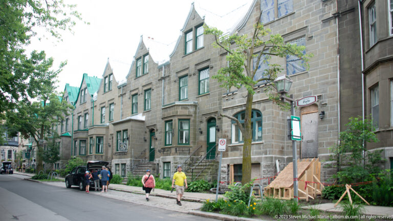 Historic stone houses with gabled roofs and bay windows line a quiet street where several pedestrians walk beside young trees.