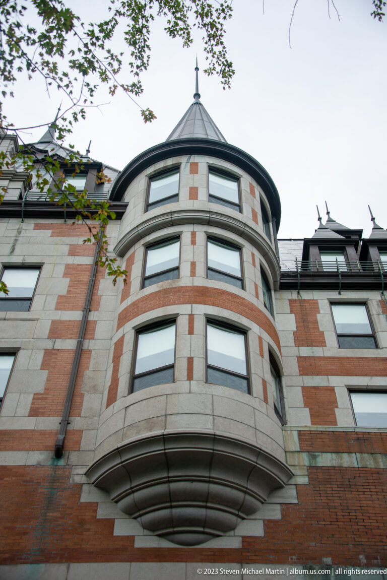 Upward view of a rounded multistory corner bay in red brick and pale stone beneath a shallow conical metal roof.