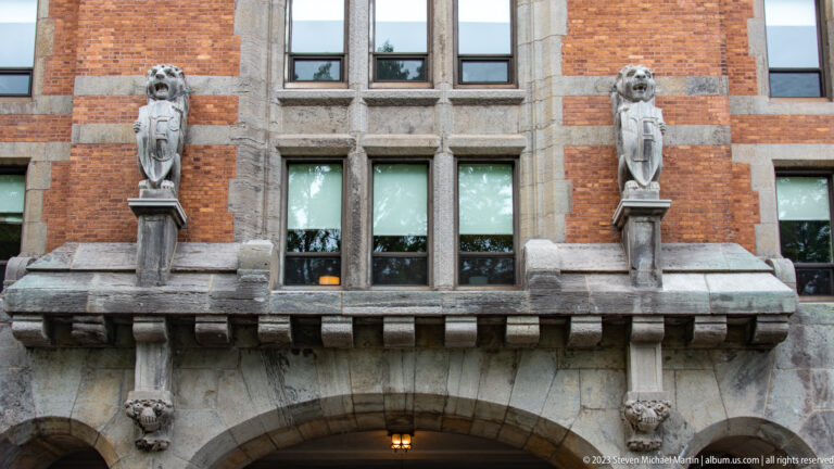 Two carved stone figures flank tall windows above a broad arch on a red-brick and stone historic façade.