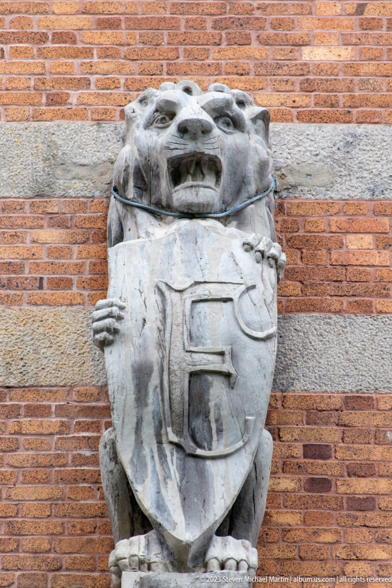 Weathered pale stone carving of grotesque figures holding a shield, mounted against a red-brick wall.