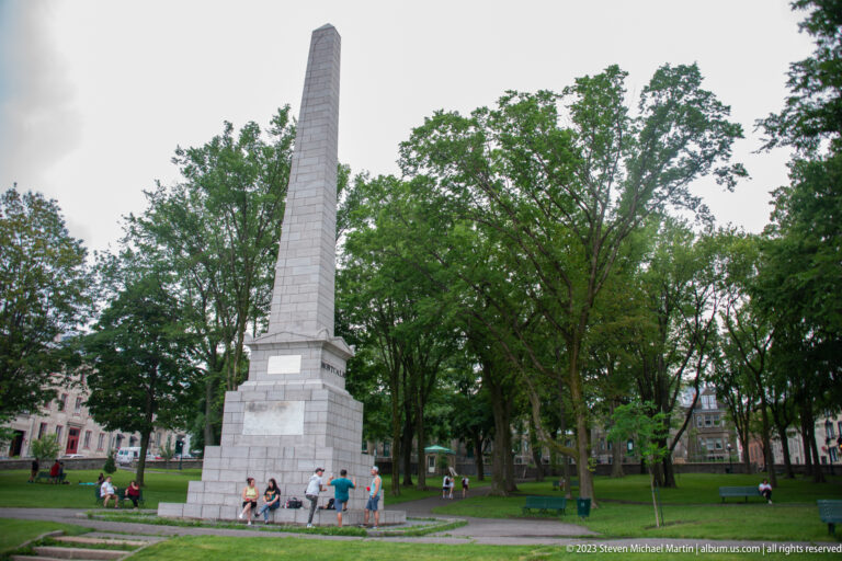 Tall pale stone obelisk of the Wolfe-Montcalm Monument rising above visitors, lawn, and mature trees in Governors Garden.
