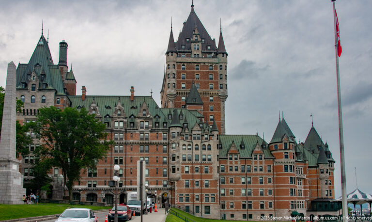 Château Frontenac with copper roofs, turrets, dormers, and tall central tower rising above cars and pedestrians.