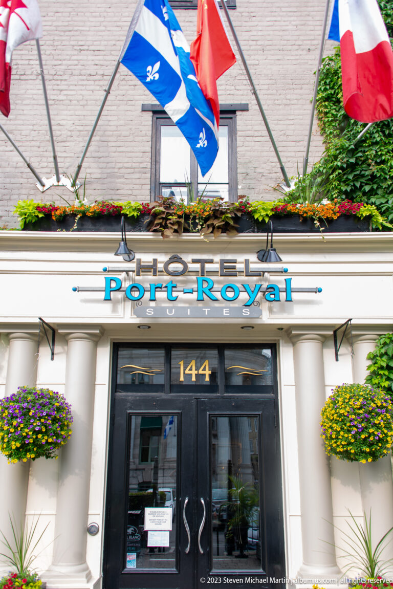 Hôtel Port-Royal entrance at number 144 with Quebec and Canadian flags, white columns, and hanging flower baskets.