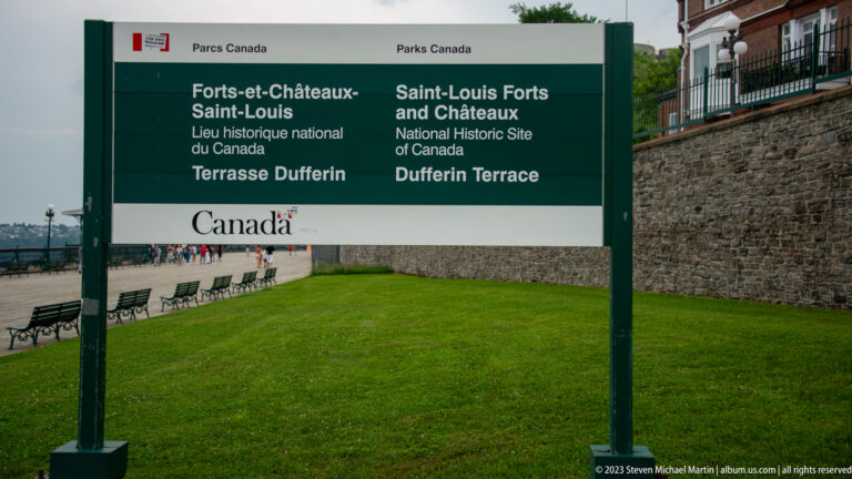 Green bilingual Parks Canada sign naming Dufferin Terrace and Saint-Louis Forts and Châteaux National Historic Site beside lawn and boardwalk.