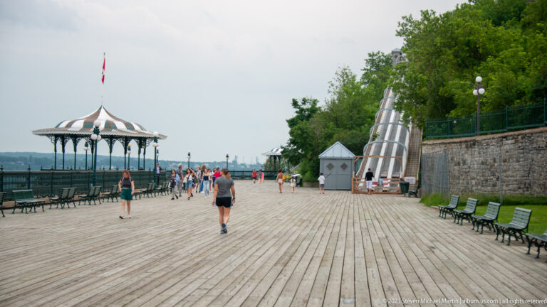 Wooden Dufferin Terrace boardwalk with pedestrians, benches, striped-roof gazebo, stone wall, and river view beyond.