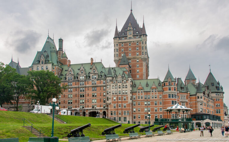Château Frontenac behind a grassy terrace lined with historic black cannons, with pedestrians near the hotel entrance.