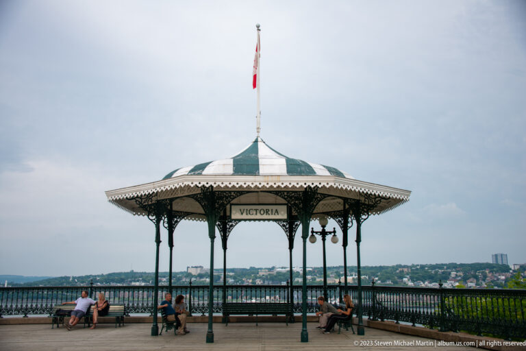 Ornate open-sided Victoria gazebo with striped roof and Canadian flag on Dufferin Terrace, overlooking the St. Lawrence River.