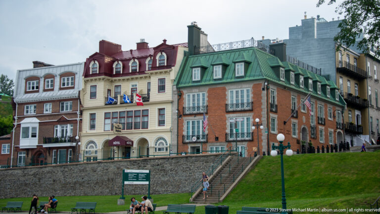 Historic brick and stucco buildings with dormered roofs and flags above a stone wall and broad stair beside Dufferin Terrace.