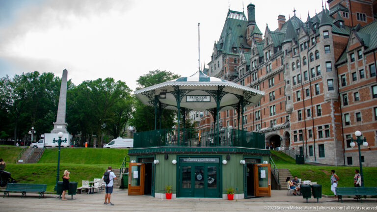 Green-and-white Dufferin Terrace kiosk and entrance with visitors in front of the towering Château Frontenac.