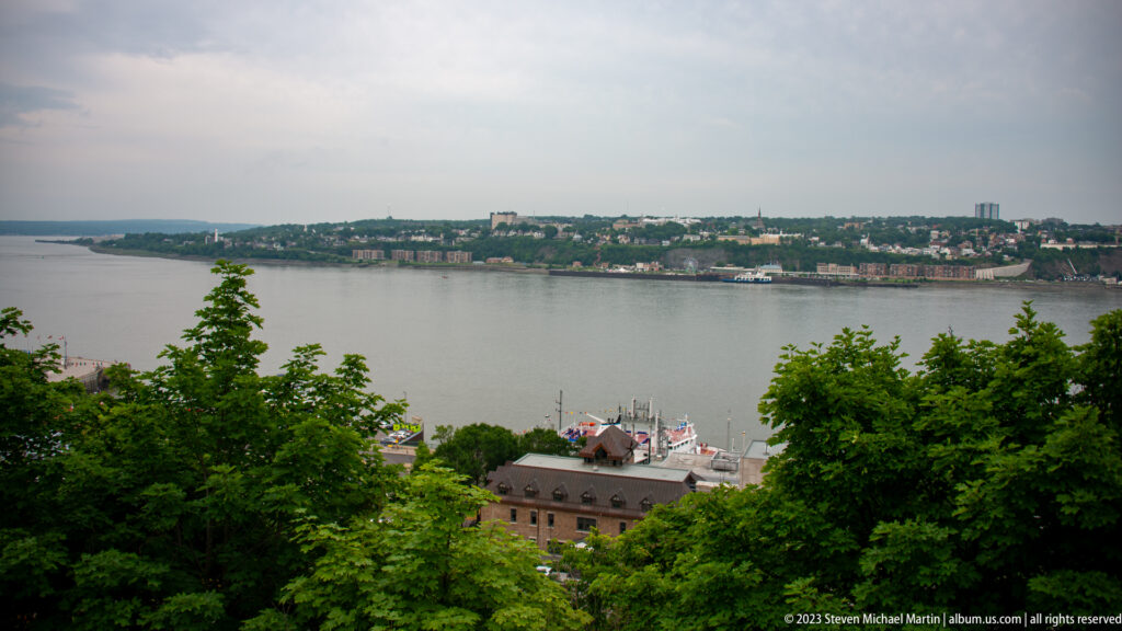 Wide view across the St. Lawrence River from a tree-lined high overlook, with rooftops below and development on the opposite shore.