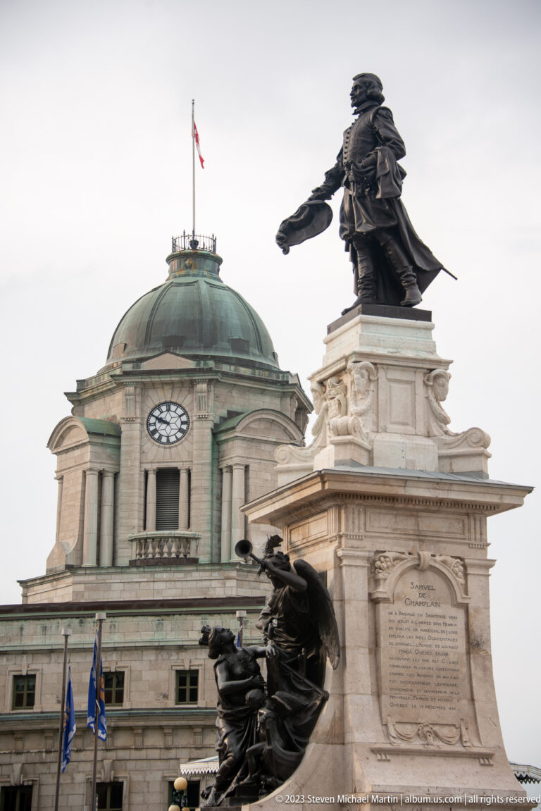 Bronze standing figure atop the Samuel de Champlain monument, with additional sculpture at the base and a domed building behind.