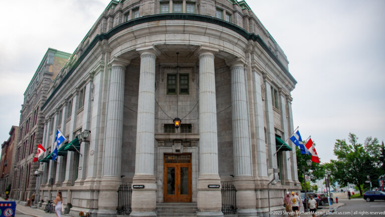 Rounded stone corner façade with tall columns, large wooden doors, flags, and pedestrians along the sidewalk.