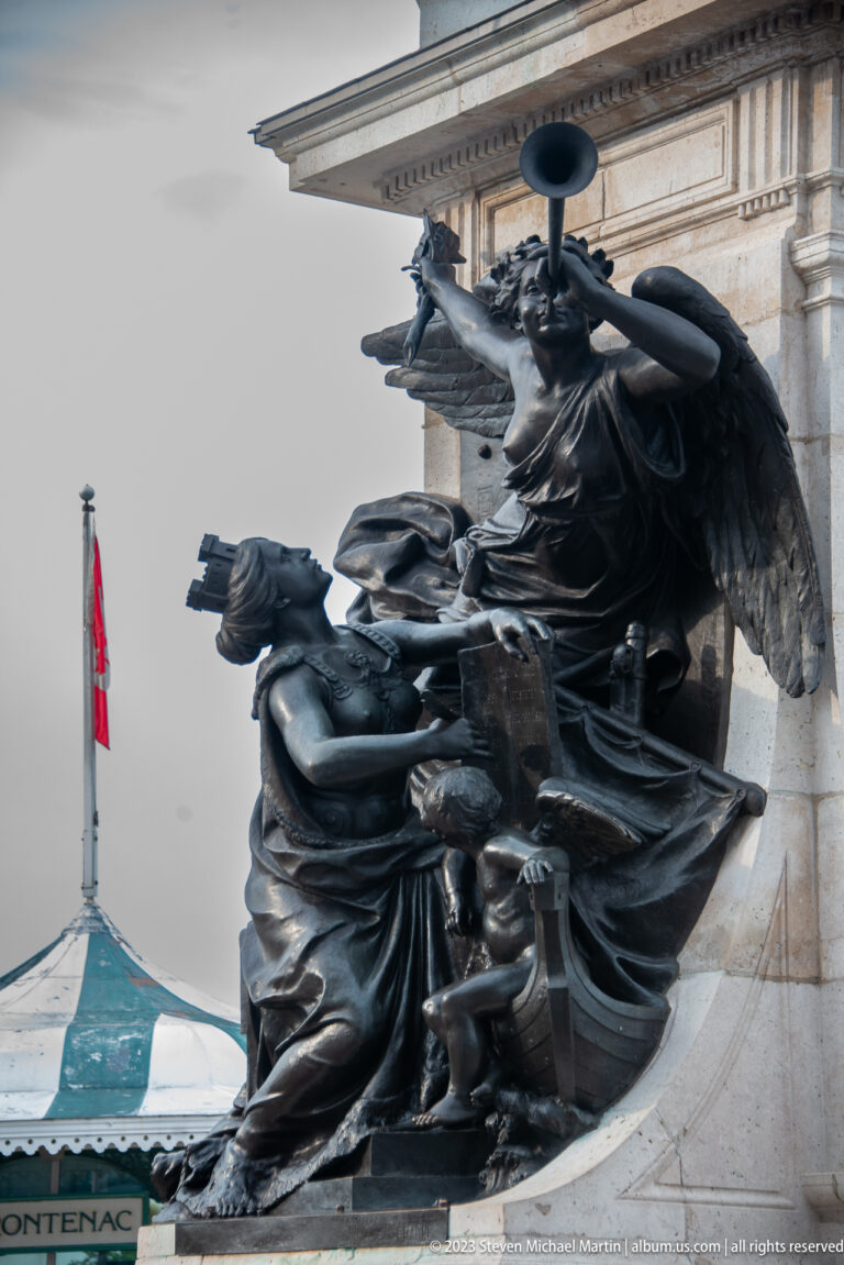 Close view of dark bronze allegorical figures, including a winged female figure, clustered against the pale stone Champlain monument.