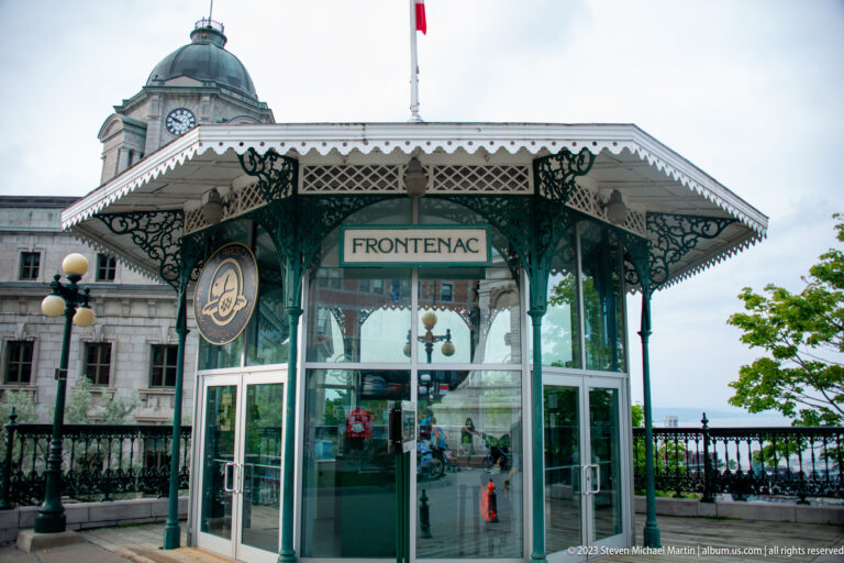 Glass-and-iron pavilion labeled FRONTENAC with decorative brackets, Canadian flag, historic stone building, and promenade railing.