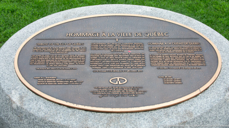 Oval bronze plaque titled Hommage à la Ville de Québec with multilingual text, set on a pale stone base beside grass.
