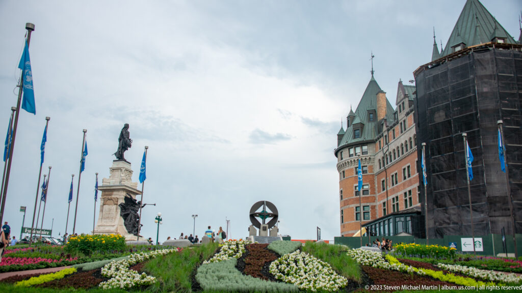 Patterned flower beds lead toward a stone monument, flags, and the turreted Château Frontenac under an overcast sky.