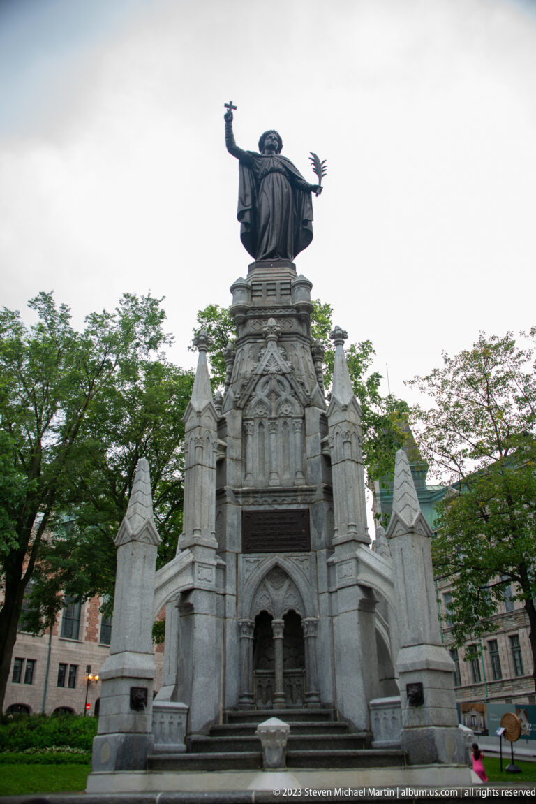 Large pale stone monument with Gothic-inspired arches and pinnacles, topped by a robed bronze figure with an outstretched arm.