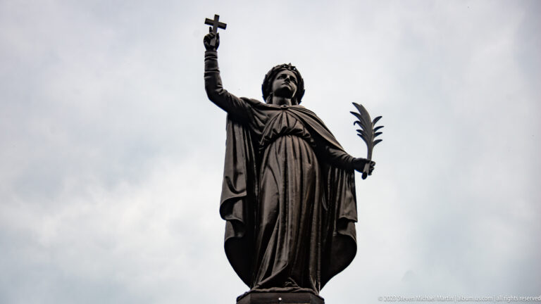 Bronze robed figure against a gray sky, raising a cross in one hand and holding a palm frond in the other.